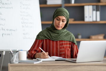 Wall Mural - Muslim teacher in headscarf sitting at desk, writing, using pc