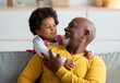 Cheerful little black girl play with older grandfather from back, hugging man at home interior