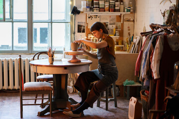 Poster - Creative ceramist molding clay vase sitting at table wearing dirty apron during master class in studio. Young businesswoman, pottery studio owner making handmade ceramics for holiday sale in workshop