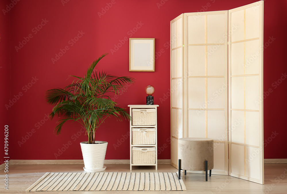 Stylish folding screen and houseplant near red wall