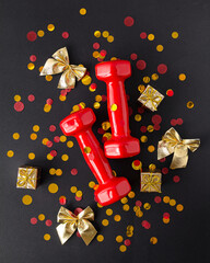 Wall Mural - Exercises, fitness and development of the concept of a merry Christmas and a happy New Year. Red dumbbells, bows and gifts with ribbons on a black background. Flat lay, top view. Vertical photography