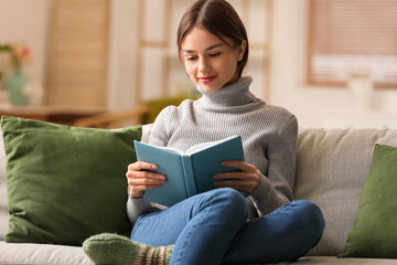 Beautiful young woman reading book on sofa at home