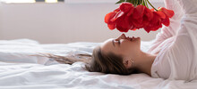 A Young Beautiful Caucasian Woman Lies On A Bed In A White Shirt Holds Red Tulips In Her Hands And Inhales Their Aroma.