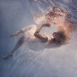 A woman with blond hair in a white dress swims underwater as if flying in zero gravity