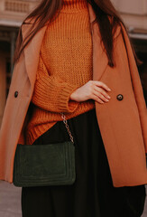 Wall Mural - Cropped female figure with green handbag in a brown cozy autumn warm coat and knitted orange sweater. Street casual autumn or winter fashion.