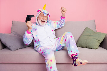 Poster - Portrait of handsome trendy cheerful grey-haired man sitting on divan playing game rejoice isolated over pink color background