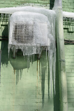 Giant Icicles Hang From The Air Conditioner On The Wall Of The Brick Green Building. Dangerous Icicles Winter Concept.