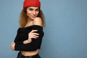 Wall Mural - Photo of beautiful self-confident sexy young brunette curly woman isolated over blue background wall wearing black crop top and red hat looking at camera