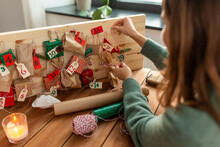 Winter Holidays, Christmas And Hobby Concept - Hands Hanging Gift With Tag To Handmade Advent Calendar At Home