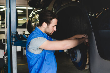 Young caucasian handsome car auto mechanic technician repairing fixing car tires wheels in special blue robe uniform at garage workshop service station.