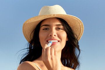 Poster - people, summer and swimwear concept - portrait of happy smiling young woman in bikini swimsuit and straw hat applying lip balm on beach