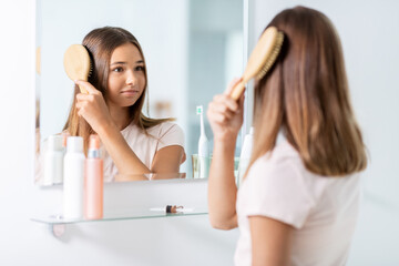 Poster - beauty and people concept - teenage girl looking to mirror and brushing hair with comb at home bathroom