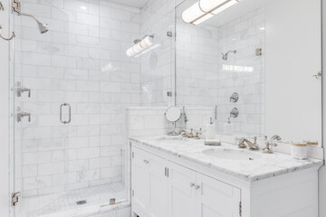 A luxurious white bathroom with a double vanity and bronze faucets, a white subway tile shower, and glass door.