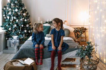 Wall Mural - two little girls are sitting on the edge of a large bed looking at each other against the background of a Christmas tree. happy christmas