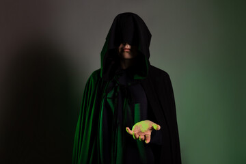 Wall Mural - A mystical character in a black hoodie stretches out his hand to you. Witch in a black hood on her face, dark background green backlight