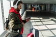 © NFstock - Side view of the relaxed young male is standing near the window and waiting for departure. He is traveling with suitcase. Guy is holding smartphone and looking at the screen with smile