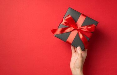 Wall Mural - First person top view photo of girl's hand holding black giftbox with red ribbon bow on isolated red background with empty space