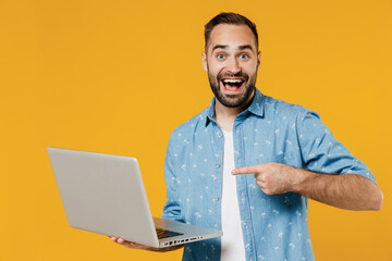 Young surprised smiling happy freelancer copywriter satisfied caucasian man 20s wear blue shirt hold use work point finger on laptop pc computer isolated on plain yellow background studio portrait