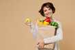 Young smiling vegetarian cheerful woman 20s in casual clothes hold paper bag with vegetables holding apple fruit eating isolated on plain pastel beige background studio portrait. Shopping concept