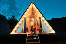 Woman Drinking Tea On The Porch Of A Wooden Lodge With Lights Of Garlands In The Evening. The Concept Of Glamping And Renting A Chalet For Weekend
