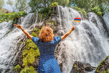 Wall Mural - Happy woman traveler with armenian flag is resting near the beautiful and large Shaki waterfall in Armenia. Fresh water and tourist attractions concept