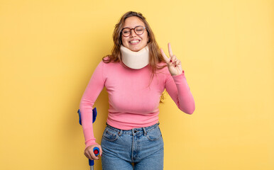 Wall Mural - hispanic pretty woman smiling and looking friendly, showing number two. crutch and collar accident