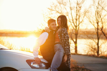 Wall Mural - Man and woman in holiday honeymoon trip. Couple is hugging in sunset near car outdoors. Lovers traveling together. Lifestyle Candid portrait. Romantic sunset journey