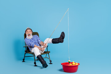 Sticker - Full size photo of happy grey haired positive cheerful old man enjoy fish hold rod sit chair isolated on blue color background