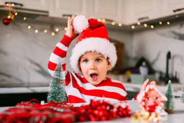 Poster - Funny toddler boy in a Santa hat is sitting in the kitchen with a decor for Christmas. Holidays, Traditions, baby food and health concept.