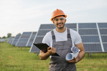 Portrait of smiling Arabian engineer in uniform, orange helmet and safety glasses holding blueprints and clipboard while standing at solar farm. Concept of people and alternative energy.