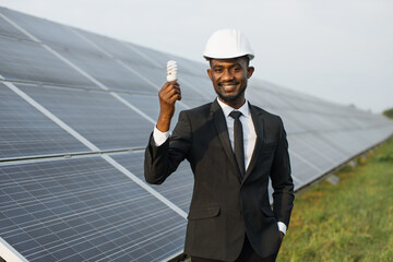 African american man with light bulb in hand standing on field with rows of solar panels. Engineer wearing black suit and white helmet. Concept of alternative energy and environment.