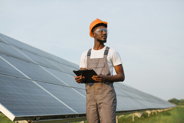 Wall Mural - Professional African American technician in helmet and glasses holding clipboard and looking away during inspection of solar station. Renewable energy and ecology concept.