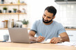© Kateryna - Positive modern Indian man, freelancer, designer or student, sitting at a kitchen at home, using laptop, studying or working online, smiling friendly, takes notes. E-learning concept