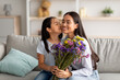 I love you, mommy. Pretty asian girl kissing her mum in cheek and giving bunch of flowers, sitting on sofa