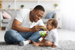 Childcare Concept. Caring Young Black Father Feeding Adorable Baby Son At Home