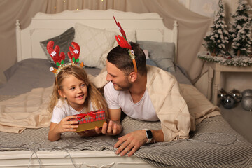 Wall Mural - a father is lying on a bed with a little daughter in a deer antler rim watching a Christmas gift box