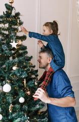 Wall Mural - Happy dad and daughter decorate the Christmas tree. A little girl is sitting on Daddy's shoulders, holding a toy on the Christmas tree. happy christmas