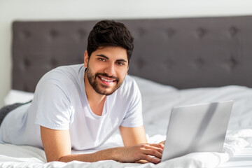 Wall Mural - Always online. Young arab man working on laptop distantly, lying on bed and smiling to camera, free space