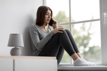 Canvas Print - Sad upset depressed european millennial woman sitting on windowsill talking on phone, getting bad news