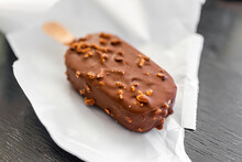 Macro Closeup Of One Chocolate Almond Nuts Ice Cream Bar On Wood Stick With Plastic White Wrap Packaging Package On Kitchen Table