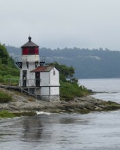 Old White Lighthouse With Red Glass Along Rocky Riverside And Against Hazy Sky