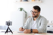 © Prostock-studio - Smiling mature caucasian guy blogger with beard in glasses make notes and look at smartphone webcam