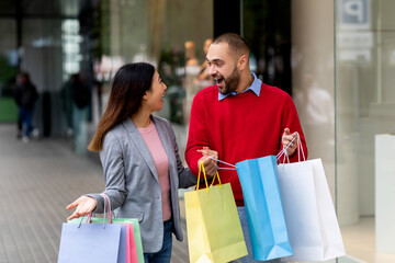 Wall Mural - Excited international couple with gift bags standing near shopping mall, shouting OMG, overjoyed about seasonal sales