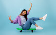 Joyful asian lady sitting on skateboard with legs up and smiling to camera, showing v-sign gesture, blue background