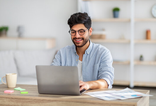 Wall Mural - Portrait of happy arab freelancer man sitting at desk with laptop computer at home office, looking and smiling at screen