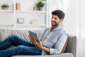 Wall Mural - Relaxed arab guy resting on sofa with digital tablet, browsing social networks or shopping online, free space