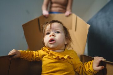 Wall Mural - One small Caucasian baby girl playing by her mother in the cardboard box at home sitting in the box real people family and growing up concept copy space