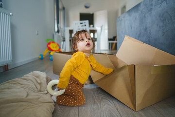 Wall Mural - One small Caucasian baby girl playing by the cardboard box at home alone