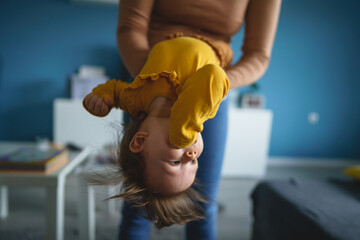 Wall Mural - One small caucasian baby girl happy in hands of her mother holding her at home in day head down having fun real people front view copy space family bonding leisure concept