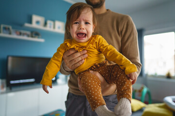 One small caucasian baby girl crying unhappy frustrated in hands of her father holding her at home in day real people front view copy space